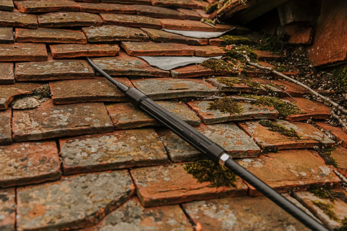 Rust-colored roof tiles with a Kerminator end tube inserted underneath them, roof showing signs of wear and moss growth.